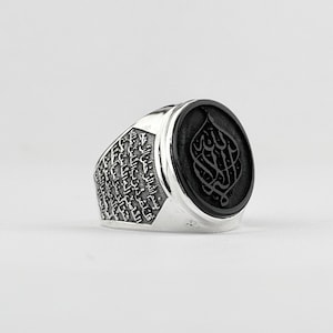 May include: A silver ring with a black onyx stone featuring Arabic calligraphy. The band of the ring is engraved with Arabic script.