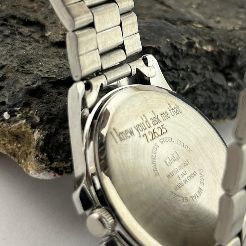 May include: Close-up of a silver stainless steel watch. The watch back has the inscription "I knew you'd ask me that 7.26.25". The watch band is made of silver metal links. The watch is water resistant.