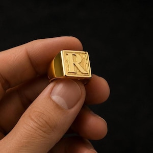May include: A gold-toned signet ring featuring a square face with a raised, embossed letter R. The surface of the square face has a textured, slightly weathered appearance, while the band and the letter R have a smooth, polished finish.