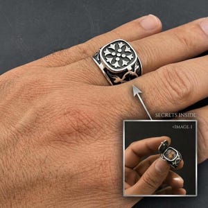 May include: A silver-toned metal ring featuring a square face with a floral-inspired cutout design. The ring includes a hinged compartment that opens to reveal a small hollow space, with the text SECRETS INSIDE and IMAGE 1 visible.