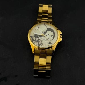 May include: A gold-toned wristwatch with a gold band and a round face. The watch face features a black and white image of a family. The watch has a classic design with a metallic finish.