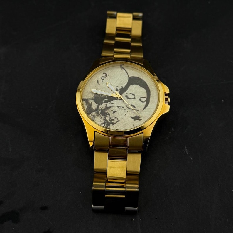 May include: A gold-toned wristwatch with a gold band and a round face. The watch face features a black and white image of a family. The watch has a classic design with a metallic finish.