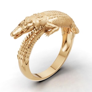 May include: A gold ring featuring a detailed, textured alligator design that wraps around the finger. The alligator's head and tail meet at the front, showcasing intricate scales and a polished finish on the band's interior and exterior surfaces.