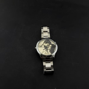 May include: A silver wristwatch with a stainless steel band. The watch face features a black and white image of a couple. The watch is a classic design, suitable for everyday wear.
