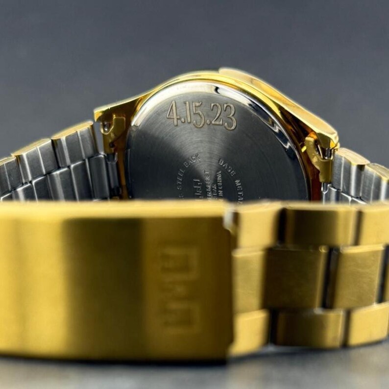 Puede incluir: Primer plano de un reloj de pulsera dorado con una correa plateada. La parte trasera del reloj muestra la fecha "4.15.23". La correa del reloj est&aacute; hecha de eslabones rectangulares.