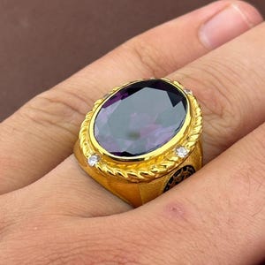 May include: A gold ring with a large, oval-cut purple gemstone. The ring has a textured gold band with three small, clear accent stones. The ring is on a finger.