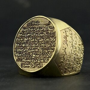 May include: A gold-toned metal ring featuring an oval face and wide band, both densely engraved with intricate Arabic calligraphy. The text is deeply etched into the surface, creating a textured, high-contrast appearance across the entire piece.