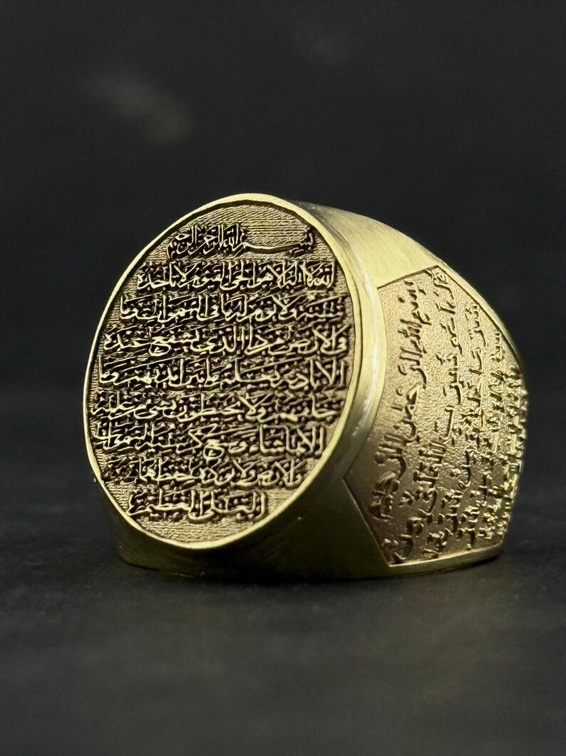 May include: A gold-toned metal ring featuring an oval face and wide band, both densely engraved with intricate Arabic calligraphy. The text is deeply etched into the surface, creating a textured, high-contrast appearance across the entire piece.