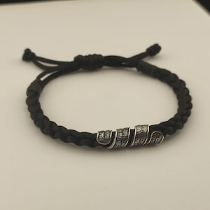 May include: A black braided cord bracelet featuring three silver-toned metal cylindrical beads. Each bead is engraved with intricate, detailed patterns. The bracelet includes an adjustable sliding knot closure with two loose cord ends.