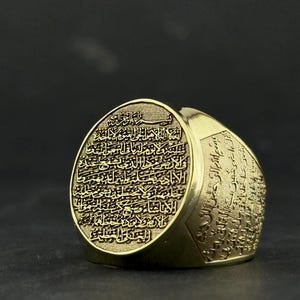 May include: A gold-toned ring featuring an oval face engraved with intricate Arabic calligraphy. The band also displays detailed script along its sides, with a textured background behind the lettering on the face, creating a high-contrast appearance.