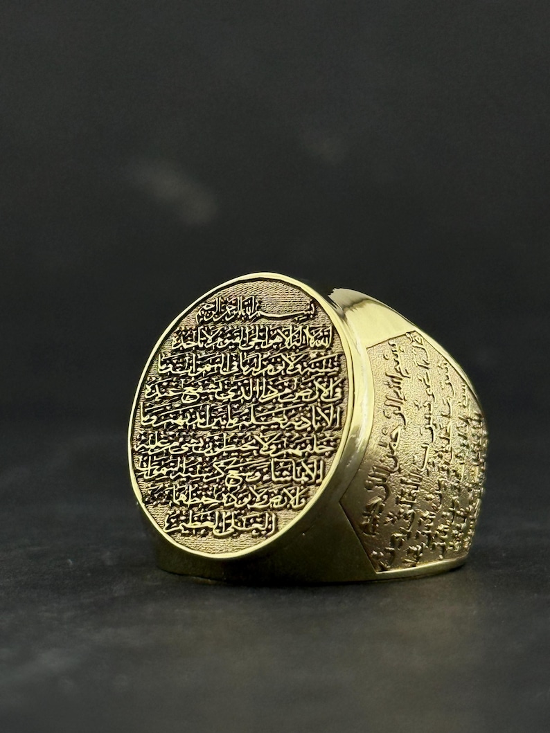May include: A gold-toned ring featuring an oval face engraved with intricate Arabic calligraphy. The band also displays detailed script along its sides, with a textured background behind the lettering on the face, creating a high-contrast appearance.