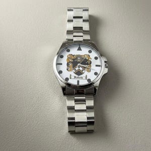 May include: A silver wristwatch with a white face featuring a family crest design. The watch has a stainless steel band and the name "Roberta" is printed on the face. The watch is a classic accessory.