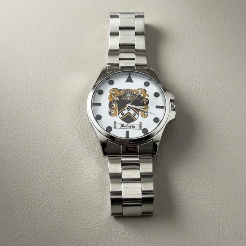 May include: A silver wristwatch with a white face featuring a family crest design. The watch has a stainless steel band and the name "Roberta" is printed on the face. The watch is a classic accessory.