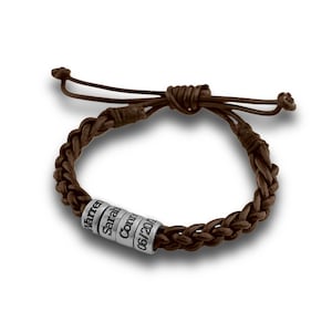 May include: A brown braided leather bracelet featuring four cylindrical silver-toned metal beads. Each bead is engraved with text, including the names Warren, Sarah, and Connor, followed by a date formatted as 06/20/12 on the final bead.