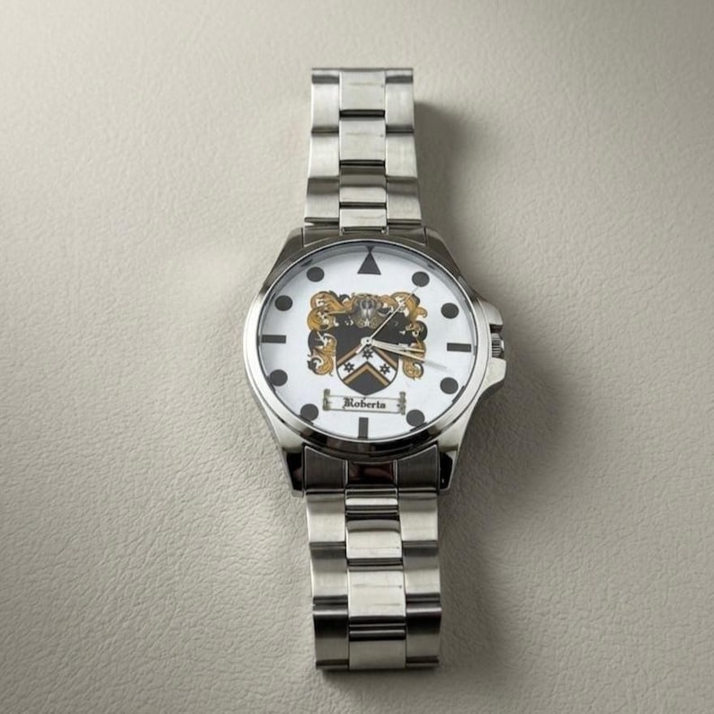 May include: A silver wristwatch with a white face featuring a family crest design. The watch has a stainless steel band and a round face with black markers and the name "Roberts" below the crest. The watch is a classic accessory.