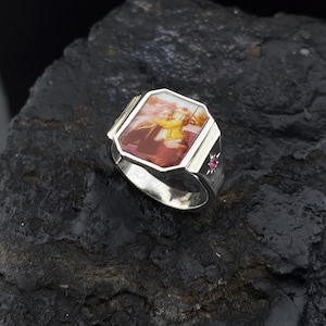 May include: A silver ring with a square-shaped bezel featuring a photo of two people. The bezel has a beveled edge and the ring has a small red gemstone on the side.