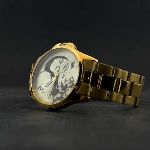 Puede incluir: Reloj de pulsera dorado con una esfera detallada con un dise&ntilde;o en blanco y negro. El reloj tiene una correa dorada y una esfera redonda. El reloj est&aacute; sobre un fondo oscuro.