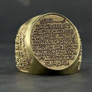 May include: A gold-toned ring featuring a large, circular face engraved with intricate Arabic calligraphy. The sides of the ring band are also decorated with detailed, embossed script patterns that wrap around the entire circumference of the metal.