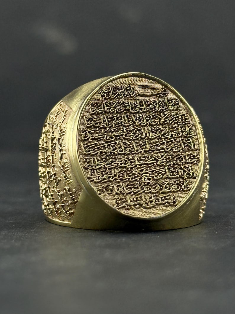 May include: A gold-toned ring featuring a large, circular face engraved with intricate Arabic calligraphy. The sides of the ring band are also decorated with detailed, embossed script patterns that wrap around the entire circumference of the metal.