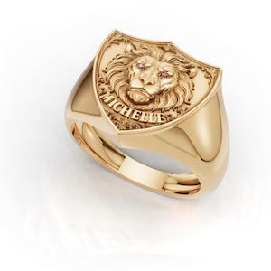 Puede incluir: Un anillo de sello de oro con una cara en forma de escudo que presenta un diseño de cabeza de león en relieve. El león tiene dos pequeñas piedras rosas como ojos. El nombre MICHELLE está grabado en letras mayúsculas bajo el león.