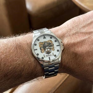May include: A silver wristwatch with a white face featuring a family crest design. The watch has a stainless steel band and a round face with black markers and the name "Roberts" displayed.