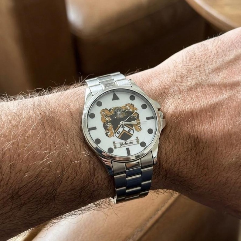 May include: A silver wristwatch with a white face featuring a family crest design. The watch has a stainless steel band and a round face with black markers and the name "Roberts" displayed.