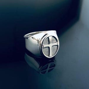 May include: A silver signet ring with a cross design. The ring has a wide band and a flat, oval face with a textured cross. The ring is highly polished and reflects the dark surface it rests upon.