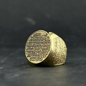 May include: A gold-toned ring featuring an oval face and wide band, both densely engraved with intricate Arabic calligraphy. The metal surface has a polished finish, and the detailed script covers the entire visible exterior of the jewelry piece.