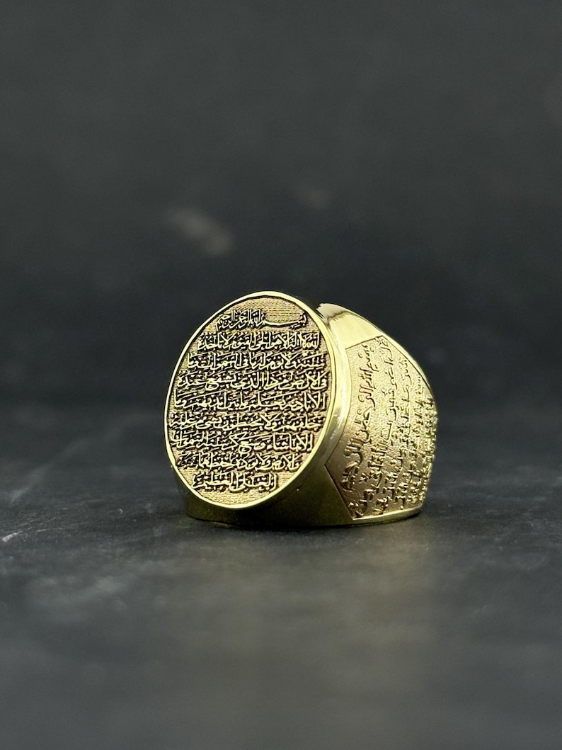 May include: A gold-toned ring featuring an oval face and wide band, both densely engraved with intricate Arabic calligraphy. The metal surface has a polished finish, and the detailed script covers the entire visible exterior of the jewelry piece.