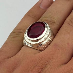Peut inclure: Une bague en argent avec une grosse pierre précieuse rouge foncé de forme ovale. La bague a un anneau texturé avec un motif en forme de corde autour du sertissage de la pierre. La bague est sur une main.