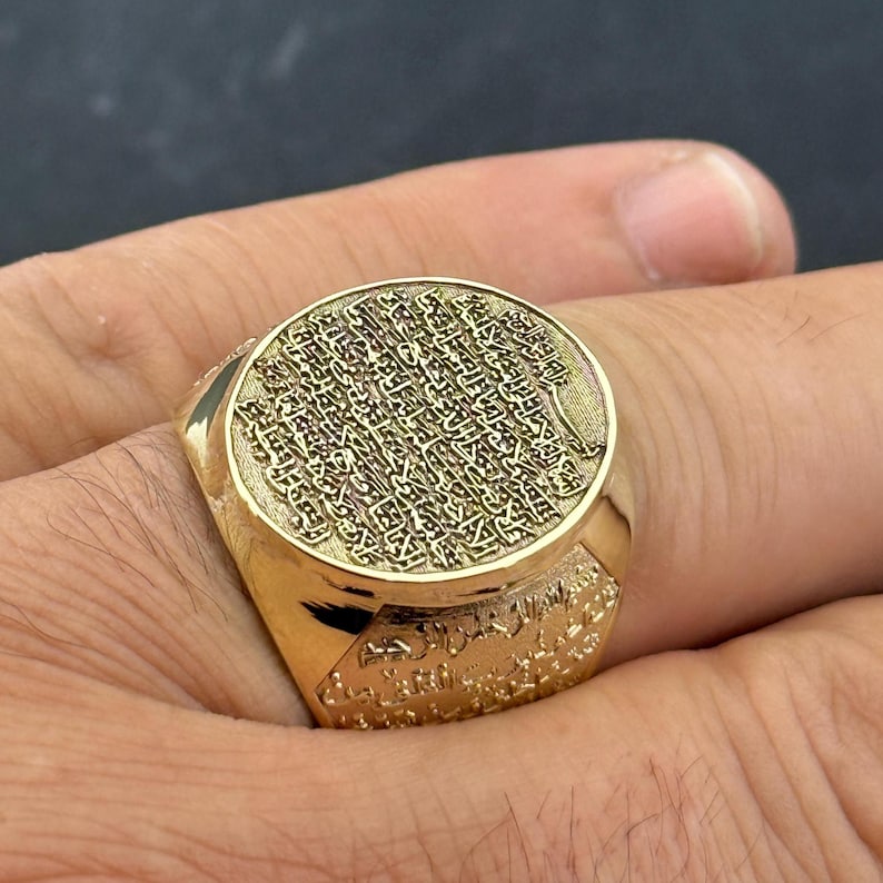 May include: A gold signet ring featuring a large, circular face engraved with intricate Arabic calligraphy. The band of the ring also displays detailed, embossed Arabic script that wraps around the sides, creating a textured, metallic finish.