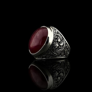 May include: A silver ring featuring a large, smooth, oval-shaped deep red gemstone set in a polished metal bezel. The band of the ring is crafted from silver and displays intricate, raised scrollwork and floral-inspired engravings throughout.