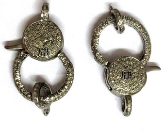 May include: Two silver-toned clasps with a round, pave-set diamond design. Each clasp has a small, round loop and a larger, round loop with a small, pointed tab. The clasps are marked with the initials "KJB".
