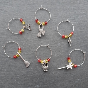 Peut inclure: Six charmes en argent pour verres à vin avec des perles rouges et jaunes. Chaque charme présente un motif différent sur le thème d'Harry Potter : un balai, un hibou, un éclair, une écharpe, une baguette avec une étoile et un triangle avec un cercle à l'intérieur.