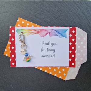 May include: A white card with the text "Thank you for being awesome!" and a rainbow design. A silver charm with a rainbow bead and wings is attached to the card.