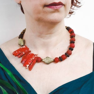 Op de afbeelding: Een statement ketting met rode en bruine kralen, met een grote, geometrische, rode stenen hanger. De ketting wordt gedragen door een persoon met een tealblauwe top.