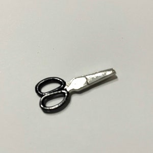 Könnte beinhalten: Eine kleine Schere mit schwarzen Griffen und einer silbernen Klinge. Die Schere liegt auf einer weißen Oberfläche. Die Klingen sind leicht geöffnet.