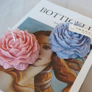 May include: Two floral shaped candles, one pink and one light blue, on a magazine with the word "Botticelli" visible. The blue candle has a small white tag that says "Thank You for your kindness".