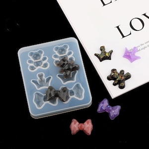 May include: A silicone mould with six cavities for making resin crafts. The cavities are shaped like bears, crowns, and bows. There are also several finished resin pieces in the image, including a purple crown, a purple bow, a black bear, and a black bow.
