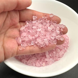 May include: A close-up of a white bowl filled with pink rose quartz chips. The chips are small and irregular in shape, and they have a translucent appearance.