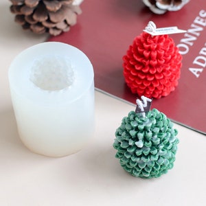 May include: A clear, cylindrical silicone candle mould, alongside two pinecone-shaped candles. One candle is red, the other is green. The candles have a textured surface, and the mould has a textured interior.