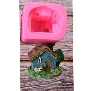 May include: Pink silicone mould for making a miniature house with a thatched roof, green walls, and a brown door. The mould is shown with a finished house made of resin or similar material.