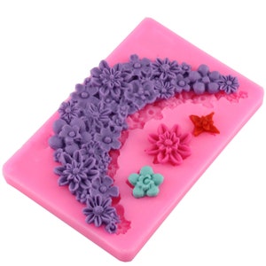 May include: Pink silicone mould with a design of a floral garland and three individual flowers. The garland is made up of purple flowers and the individual flowers are purple, red, and blue.