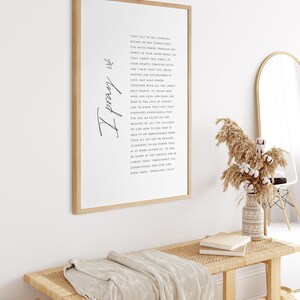 May include: A framed print with a white background and black text. The text is a prayer about the love of Christ and the power of God. The print is hanging on a white wall above a wooden bench with a woven seat.