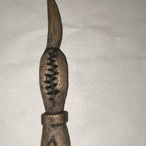 Può includere: Una scultura in legno di una figura stilizzata con una testa appuntita e un motivo intagliato sul corpo. La figura è di colore marrone e ha una piccola base triangolare.