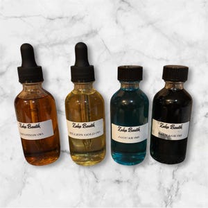 May include: Four glass bottles with black droppers and labels. The bottles contain liquids in shades of amber, yellow, blue, and dark brown. The labels read "Zaka Boutik" and include the names "DEVOTION (W)", "MILLION GOLD (W)", "JAGUAR (OD)", and "SAUVAGE (M)".