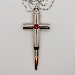 May include: A silver-colored cross pendant necklace. The cross is made from bullet casings and features a red gemstone at the center. The necklace has a ball chain.