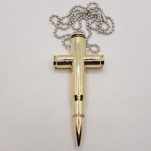 May include: A gold-colored bullet casing cross pendant on a silver chain. The cross is made from a single bullet casing, with the bullet tip still attached.