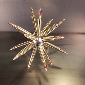 May include: A decorative metal sculpture resembling a starburst. The sculpture is made of brass-colored bullet casings radiating from a central silver sphere. The tips of the bullets are copper-colored.
