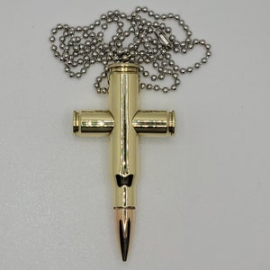 May include: A gold-colored bullet casing cross pendant on a silver chain. The cross is made from four bullet casings, with the bullet tip at the bottom.